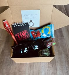 an open cardboard box with sunglasses, notepad and other items in it on a wooden floor