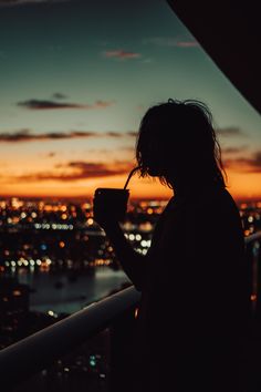 a person standing on top of a building drinking from a cup at night with the city lights in the background