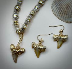 Actual shark tooth earrings and necklace set in a one-of-a-kind handmade design. The shark teeth were found locally along the Carolina coasts and wire wrapped using gold-colored copper wire and hypoallergenic findings. The shark tooth pendant has gold foil accents along the top and is on a handmade twine cord with puka shells and gold accents woven into the cord. The cord is 18 to 21 inches long. The pendant shark tooth is about 1 inch high and 0.75 inches wide. The shark teeth in the earrings a Shark Tooth Earrings, Tooth Earrings, Shark Tooth Pendant, Tooth Pendant, Earrings And Necklace Set, Puka Shell, Shark Tooth, Earrings And Necklace, The Shark