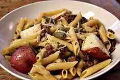 a white bowl filled with pasta and meat next to a red potato on top of a wooden table