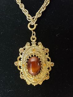 This pendant necklace has an ornate gold tone embossed frame with prong set brown stones. At the center is a huge brown cabochon and on the backside it has a hallmark of "Celebrity N.Y.". The pendant is strung on a triple ring rope chain. All stones appear to be intact with non-missing. The chain appears slightly worn due to chain and wear. This pendant necklace came from an estate sale this year. It will arrive in a gift box with FREE SHIPPING. Rope chain measures: Pendant measures: Material: Hallmark: Celebrity N.Y. Gift box FREE SHIPPING Luxury Vintage Brown Necklaces, Ornate Gold Oval Cabochon Jewelry, Antique Gold Oval Cabochon Necklace, Antique Gold Necklace With Oval Cabochon, Gold Oval Pendant Necklaces With Cabochon, Gold Necklaces With Oval Pendant Cabochon, Gold Oval Pendant Necklace With Cabochon, Gold Necklaces With Oval Cabochon Pendant, Brown Metal Pendant Jewelry