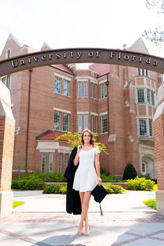 a woman in a white dress and black jacket is walking under the university of florida arch