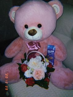 a pink teddy bear sitting on top of a chair holding a basket of flowers and a chocolate bar