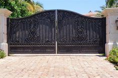 an iron gate in front of a brick driveway with palm trees and bushes behind it