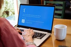 an older woman sitting at a table with a laptop computer on her lap and coffee mug in front of her