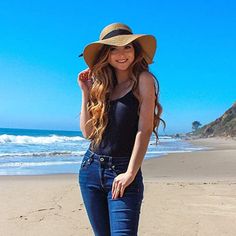 a beautiful young woman standing on top of a beach next to the ocean wearing a hat