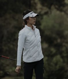 a woman in white jacket and black pants holding a golf club with trees in the background