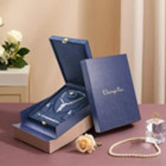 an open blue box sitting on top of a table next to a white rose and necklace