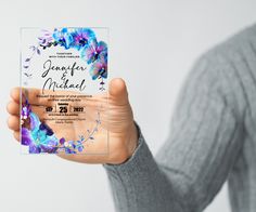 a person holding up a purple and blue wedding card with flowers on the front in their hand