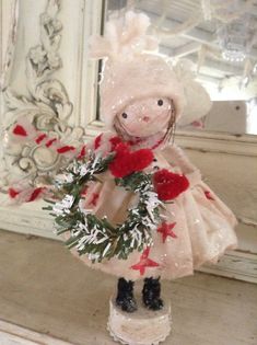 a doll holding a wreath on top of a wooden table in front of a mirror