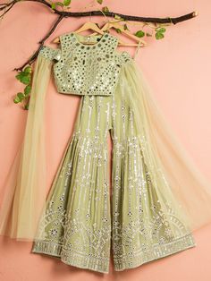 A three-piece Sage Green Mirror Sharara set from the Anisha Shetty collection. This beautiful sage green cold-shoulder blouse in rawsilk fabric is paired with a georgette sharara pant. The sage green color with mirror embroidery and mirror lace enhance this sharara set. The outfit is completed with a green dupatta in net material attached to the blouse. Green Combination Outfit Indian, Trendy Sharara Designs, Blouse Sharara Set, Green Color Sharara Suit, Fitted Pista Green Sharara With Unstitched Blouse, Sage Green Indian Outfit, Floor-length Pista Green Sharara For Navratri, Pista Green Sharara With Mirror Work For Reception, Sage Green Sharara