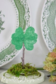 a small green tree sitting on top of a white plate next to plates and flowers