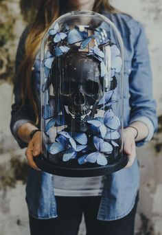 a woman holding up a glass case with blue flowers on it and a skull in the middle