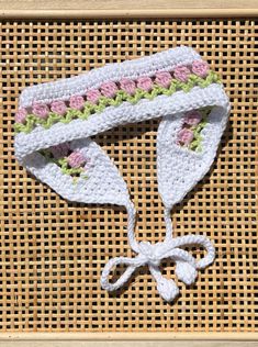 a crocheted white hat with pink and green flowers on the side, tied to a wicker board