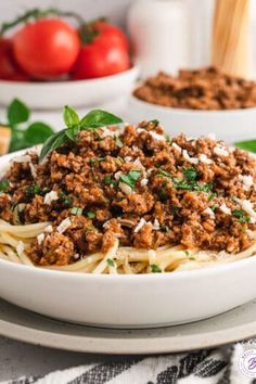 spaghetti with meat and cheese in a white bowl on a plate next to tomatoes, basil leaves and breadcrumbs