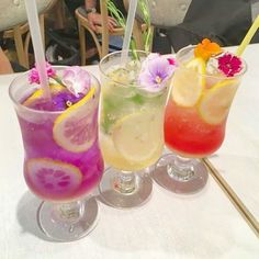 three glasses filled with different colored drinks on top of a white tablecloth covered table