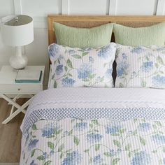 a white bed with blue flowers on it and green pillows in front of the headboard