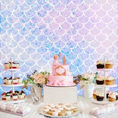 a table topped with cakes and cupcakes under a mermaid tail wallpapered backdrop