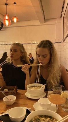 two women sitting at a table with chopsticks in their mouths, eating noodles