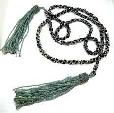 This lariat necklace is comprised of black and silver peanut beads and aqua seed beads in a spiral herringbone pattern rope, with tassels on each end. The necklace is woven with FireLine thread. Design by Linda Newberry.55.5" end to end length rope is approx. .25" wide Adjustable Lariat Beaded Necklaces With Tassels, Beaded Lariat Tassel Necklace, Adjustable Black Beaded Lariat Necklace, Adjustable Lariat Beaded Necklace With Black Beads, Adjustable Black Beads Lariat Beaded Necklaces, Thread Design, Rope Necklace, Herringbone Pattern, Lariat Necklace