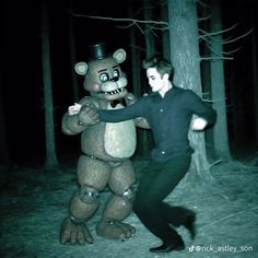 a man standing next to a giant teddy bear in the woods at night with his arm outstretched