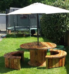 a wooden table and bench with an umbrella over it in the grass next to some bushes