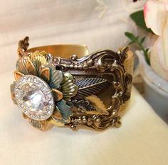 "SALE, Victorian Cuff,Vintage Crystal Button, Brass Frame,Leaf and Flowers,Large brass Cuff,Ornate Cuff,Wide Cuff,MockiDesigns,Gift Wrapped Was 64. SALE brass Cuff with vintage crystal button. An American tooled brass cuff with detailed design has a large rectangular ornate brass frame on which I places several leaf/floral findings and finally a vintage very sparkly crystal button. Under the flower I also placed different leaves. The cuff width is 1\", 2.54cm and the center plaque slightly large Framed Leaves, Brass Cuff, Crystal Buttons, Steampunk Design, Wide Cuff, Fun Fashion, Brass Frame, Vintage Crystal, Handmade Art