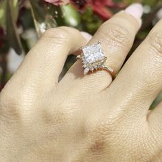 a woman's hand with a diamond ring on top of her finger and flowers in the background