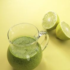 a glass pitcher filled with green liquid next to two limes on a yellow surface