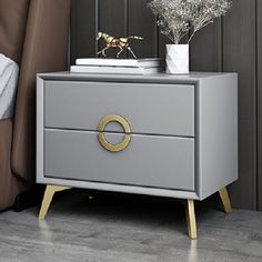 a nightstand with two drawers and a gold ring on the bottom drawer, next to a bed