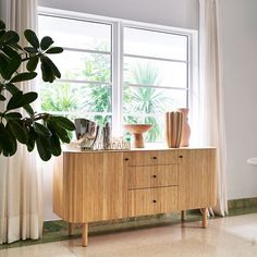 a wooden cabinet sitting in front of a window next to a plant and vases