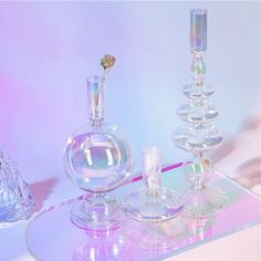 three glass vases and one candle holder on a mirrored surface with pink lighting in the background