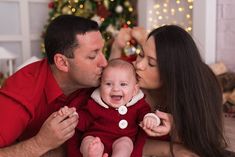 a man and woman kissing a baby in front of a christmas tree