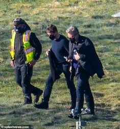 three men walking in the grass with one man wearing a safety vest and another holding a skateboard