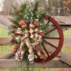 a christmas wreath is hanging on a wooden wagon wheel in front of a fence with evergreens and pine cones