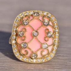 The 154th Avenue: Pink Coral And Natural Diamond Cocktail Ring In Yellow Gold. The Ring's Focal Point Is A Central Round-Cut Natural Diamond Ensconced Within A Lattice Of Yellow Gold Lace Adorned With Natural Diamonds. Below This Intricate Design Rests A Cushion Buff Top Made Of Exquisite Pink Coral. Encircling The Coral Is A Halo Of Round-Cut Natural Diamonds. The Split Shank Of The Ring Is Also Adorned With Round Brilliant-Cut Natural Diamonds. Delicate Milgrain Detailing Graces The Lacework, Both Outside The Halo And Around The Bezels. Crafted In 14 Karat Yellow Gold, The Ring Is Currently Sized At 6.75 But Can Be Resized To Fit Any Finger Size For An Additional Charge Upon Request. Yellow Gold Color, Diamond Cocktail Ring, Diamond Cocktail Rings, Gold Lace, Split Shank, Pink Coral, Intricate Design, Cocktail Ring, Cocktail Rings