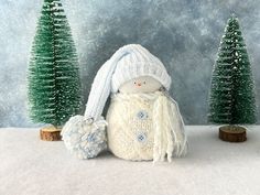 a small snowman with a white hat and scarf next to some trees in the snow