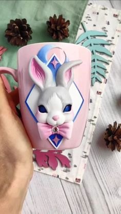 a person is holding a pink mug with a bunny face on it and pine cones in the background