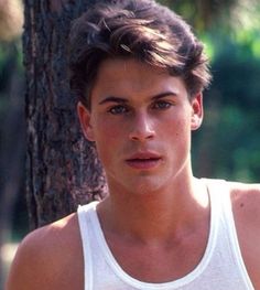 a young man standing in front of a tree wearing a white tank top and looking at the camera