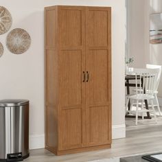 a tall wooden cabinet next to a trash can in a room with white walls and wood flooring