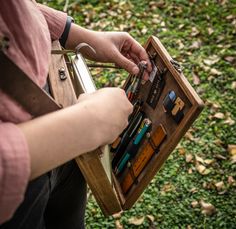 a person holding an open wooden box filled with pens and pencils on top of green grass