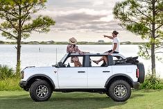 two men sitting in the back of a white truck on grass next to trees and water