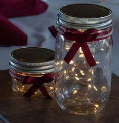 two mason jars with lights in them are sitting on a wooden table next to a red ribbon