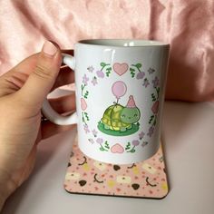 a hand holding a coffee mug with a turtle on it and pink hearts in the background