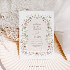 a wedding card with flowers and leaves on it next to some white feathers in the background