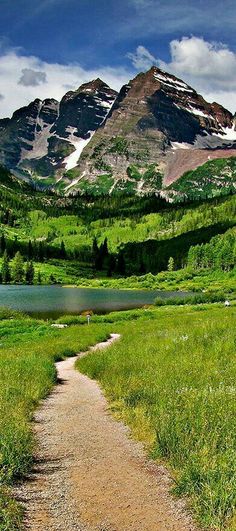 a dirt path leading to a lake in the middle of a green field with mountains behind it