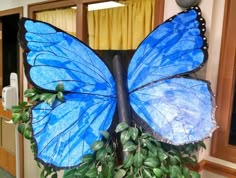 a large blue butterfly sitting on top of a green plant in front of a window