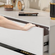 a person's hand is opening a drawer in a white cabinet with gold accents