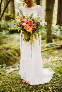 a woman standing in the woods holding a bouquet of flowers and greenery on her wedding day