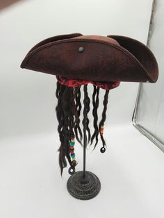 Complete your Pirate Costume this season with this Deluxe Child Caribbean Pirate Hat with Attached Dread Locs Wig. Perfect for Halloween, Cosplay, Theater and more! Featuring a tri-corne hat with attached brown faux braided hair with beads accented by a red band with floral filigree. Great for children of all ages! Tricorner hat has sweatband inside for a customized fit. Fits approximately 48 cm to 55 cm. Dreads hand approximately  long. Nicky Bigs Novelties brand is exclusively brought to you by Marsino's, a name with over 100 years of business. For Ages 3 and Older with CPSIA safety testing. Deluxe child size Caribbean pirate hat with dreadlocks, gold earring and black eye patch set. Featuring a Buccaneer tri-corne hat with attached brown faux braided hair with beads accented by a red ba Adjustable Themed Costume Hats And Headpieces, Red Themed Adjustable Costume Accessories, Adjustable Pirate Hat For Costumes, Adjustable Pirate Hat For Costume, Adjustable Pirate Costume Accessories For Costume Party, Adjustable Pirate Costume Accessories For Halloween, Pirate Costume Accessories For Costume Party, Adjustable Novelty Mini Hat For Cosplay, Adjustable Pirate Hat For Themed Events